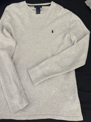 Ljusgrå långärmad tröja Polo Ralph Lauren - Ljusgrå långärmad tröja från Polo Ralph Lauren med diskret struktur och klassisk rund hals. Tröjan har en liten broderad logga på bröstet och är tillverkad i mjukt material som känns skönt mot huden. Perfekt för en clean och stilren look.Den är perfekt till vår och höst och den är i bra skick.