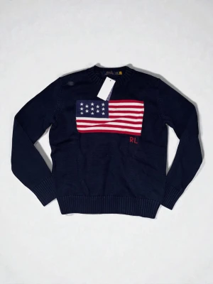 Mörkblå stickad tröja Polo Ralph Lauren - Mörkblå stickad tröja från Polo Ralph Lauren med amerikansk flagga i rött, vitt och blått på bröstet samt RL-broderi. Tröjan har rund halsringning och ribbade muddar vid ärmslut och nederkant. Perfekt för dig som gillar klassisk preppy stil.