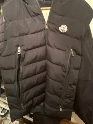 Svart dunväst från Moncler - Säljer en svart dunväst från Moncler med två stora dragkedjeförsedda fickor framtill och klassisk Moncler-logga på bröstet. Västen har quiltad design och är tillverkad i ett glansigt material som ger en exklusiv känsla. Perfekt för lager-på-lager och streetstyle.