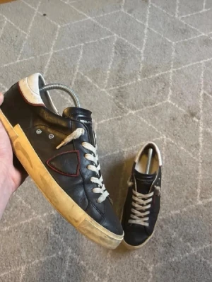 Svarta sneakers från Philippe Model - Snygga svarta sneakers från Philippe Model med vita snören, beige sula och röd detalj på sidan. Skorna har en låg profil och är tillverkade i läder med klassisk design och logotyp på tungan. Perfekta för en avslappnad streetwear-look.