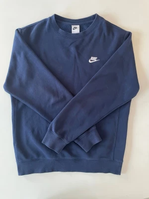 Mörkblå sweatshirt från Nike - Klassisk mörkblå sweatshirt från Nike med broderad vit logga på bröstet. Tröjan har rund halsringning, långa ärmar och ribbade muddar vid ärmslut och nederkant. Perfekt för en avslappnad och sportig stil.