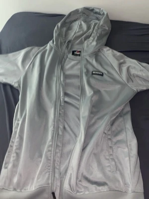 Silverfärgad hoodie från McKenzie - Snygg silverfärgad hoodie från McKenzie med dragkedja framtill och huva. Tillverkad i ett glansigt, mjukt material som ger en sportig vibe. Märkeslogga på bröstet och praktiska fickor på sidorna. Perfekt för dig som gillar streetwear och vill sticka ut.