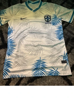 Brasil fotbollströja med palmblad - Snygg Brasilien fotbollströja från Nike i vitt med blå och grå palmbladsmönster. Tröjan har V-ringning med blå kant och Brasil-logga på bröstet. Perfekt för dig som vill sticka ut på planen eller heja på landslaget.