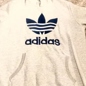 Grå hoodie från Adidas - Säljer en stilren grå hoodie från Adidas med den klassiska loggan i blått på bröstet. Tröjan har en känguruficka och långa ärmar med ribbade muddar. Perfekt för en avslappnad stil.