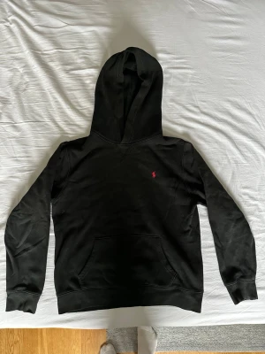 Svart hoodie från Ralph Lauren - Svart hoodie från Ralph Lauren med klassisk huva och känguruficka. Liten röd broderad logga på bröstet. Mjuk bomullskänsla och ribbade muddar vid ärmslut och nederkant. Jag är 175 cm lång och passar mig bra. Proset kan diskuteras.