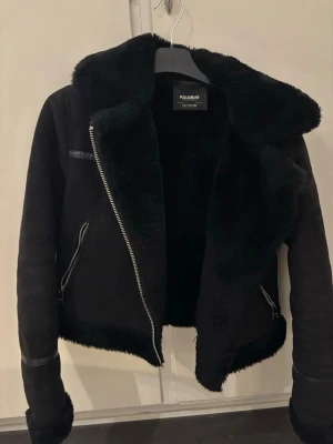 Svart skinnjacka från Pull&Bear - Snygg svart skinnjacka från Pull&Bear med mjukt foder och bred krage. Jackan har dragkedja i sned modell, två fickor med dragkedja och detaljer i fuskskinn. Perfekt för dig som vill ha en trendig och varm jacka med cool look.