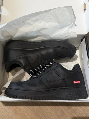 Nike Air Force 1 x Supreme svart - Svarta Nike Air Force 1 Low i samarbete med Supreme. Skorna har en clean svart läderöverdel, svarta sulor och Supreme-loggan i rött på sidan. Klassisk siluett med svarta och vita skosnören och diskreta detaljer. Använda 5 gånger, båda skosnören följer med.