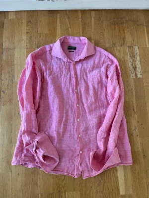 Rosa linneakjorta - Säljer en snygg rosa skjorta i linne. Skjortan har klassisk krage, vita knappar framtill och lång ärm. Perfekt för en avslappnad men ändå stilren look köpt på Åhléns för 1100.