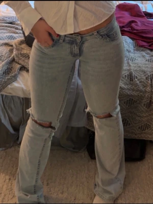 Ljusblå slitna bootcutjeans - Säljer ett par ljusblå bootcutjeans med slitningar och hål vid knäna. Jeansen har klassisk femficksdesign och broderade detaljer på bakfickorna. Perfekta för en avslappnad och trendig look. slitningar längst ner på benet. pris kan diskuteras 