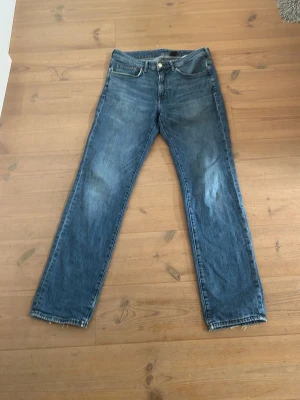 Blå raka jeans med slitna detaljer - Säljer ett par blå jeans med rak passform och slitna detaljer vid benslut och lår. Jeansen har klassisk femficksdesign och knappgylf. Perfekta för en avslappnad och trendig look.