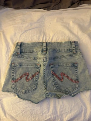 Ljusblå jeansshorts från Nelly - Säljer ett par ljusblå jeansshorts från Nelly med råa kanter och dubbla knappar framtill. Shortsen har hög midja och bakfickor med röda broderade zigzag-detaljer. Perfekta för varma dagar och en avslappnad look.
