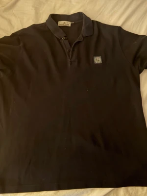 Svart pikétröja från Stone Island - Klassisk svart pikétröja från Stone Island med liten logotyp på bröstet. Tröjan har krage, knappar vid halsen och är tillverkad i mjuk bomull. Perfekt för en stilren och avslappnad look.Storlek är XXL men passar XL också! (Vissa L passar också!)
