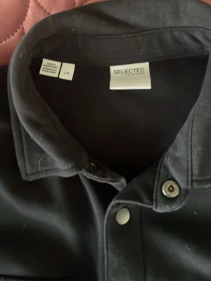 Svart overshirt med tryckknappar - Stilren svart overshirt med två stora bröstfickor med lock och tryckknappar framtill. Klassisk krage och rak passform. Perfekt att slänga över en hoodie eller t-shirt för en cool look.