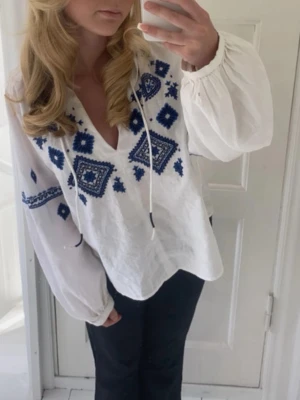 Vit blus med blå broderier - Säljer en vit boho-inspirerad blus med blå broderade mönster och knytband vid halsen. Blusen har en avslappnad passform, djup v-ringning och vida långa ärmar med resår vid handlederna. Perfekt för en trendig och avslappnad stil. Storlek xs passa mig som bruka ha xs-S