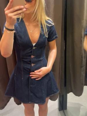 Mörkblå jeansklänning med knappar - Snygg mörkblå jeansklänning med v-ringning och korta ärmar. Klänningen har markerad midja, dekorativa sömmar och tre stora knappar framtill. Perfekt för dig som gillar en trendig och avslappnad stil.