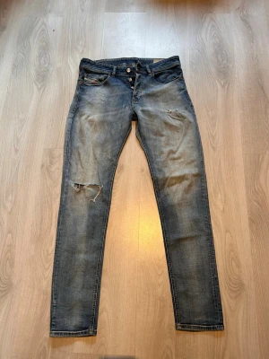 Diesel jeans - Säljer ett par blå skinny jeans med slitningar och hål på vänster knä. Jeansen har klassisk femficksmodell, knappgylf och en tvättad look med ljusare partier. Perfekta för en avslappnad och trendig stil. W30 L32