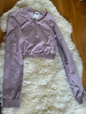 Ljuslila cropped tröja från Gymshark - Cropped långärmad tröja från Gymshark i ljuslila nyans. Tröjan har rund halsringning, ribbade muddar och tryckt logga på sidan. Tillverkad i mjukt och stretchigt material, perfekt för träning eller chill.