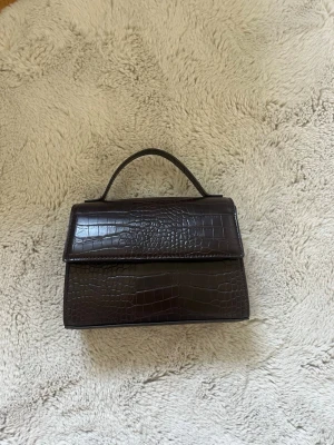 Mörkbrun croco handväska - Snygg mörkbrun handväska i croco-mönstrat konstläder med stilren, fyrkantig form och ett kort handtag upptill. Perfekt accessoar för att lyfta din outfit med en trendig touch.