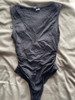 Svart body med spetsdetaljer - Snygg svart body med djup V-ringning och spetskant längs urringningen. Ärmlös modell i mjukt stretchigt material som sitter tight och framhäver figuren. Perfekt att styla med kjol eller jeans🥰