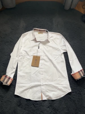 Vit skjorta från Burberry med rutiga detaljer - Snygg vit skjorta från Burberry med klassiska rutiga detaljer på insidan av ärmsluten och kragen. Skjortan har knappar framtill och en broderad logga på bröstet. Perfekt för dig som gillar stilrena plagg med en twist.