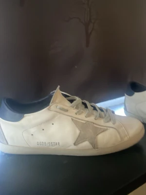 Vita Golden Goose sneakers med stjärna - Snygga vita sneakers från Golden Goose med grå stjärndetalj på sidan och svart häl. Skorna har klassisk låg profil, vita skosnören och GGDB/SSTAR-tryck på sidan. Perfekta för en trendig och avslappnad streetstyle-look.