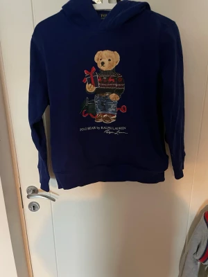 Blå hoodie Polo Bear Ralph Lauren - Säljer en blå hoodie från Ralph Lauren med den ikoniska Polo Bear-figuren på bröstet. Hoodien har huva och tryck i flera färger, där björnen bär en stickad tröja och jeans. Perfekt för dig som gillar klassisk streetwear med en twist. ÖPPEN FÖR BYTE 