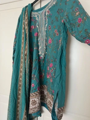 Turkos panjabi med broderi och sjal - Säljer en turkos panjabi med matchande sjal. Plagget har vackra broderier i vitt och rosa samt blommönster och dekorativa detaljer längs kanterna. Sjalen har guldfärgade mönster och matchar perfekt till kurta-setet. Perfekt för dig som gillar färgstarka och unika plagg.