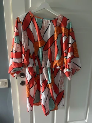 Färgglad kort omlottklänning m inbyggda shorts - Säljer en kort omlottklänning med färgstarkt abstrakt mönster i rött, orange, turkost och vitt. Klänningen har vida ärmar, knyt i midjan och v-ringning. Perfekt för dig som gillar unika plagg med mycket färg och mönster.