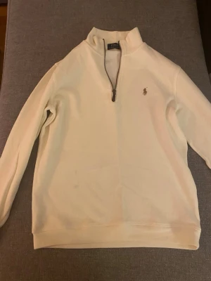 Vit halvzip från Polo Ralph Lauren - Klassisk vit tröja med halv dragkedja från Polo Ralph Lauren. Tröjan har ribbade muddar, hög krage och den ikoniska broderade loggan på bröstet. Perfekt för en clean och stilren look. Materialet känns mjukt och bekvämt.