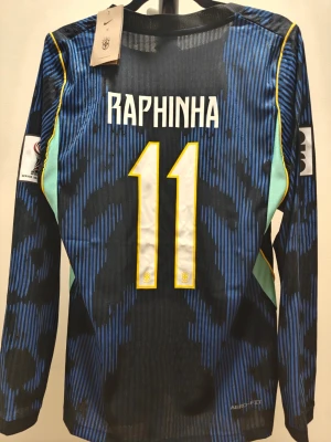 Brasilien spelar version långärmad matchtröja Raphinha - Snygg spelar version långärmad Brasilien-tröja från Nike med Raphinha och nummer 11. Tröjan är blå och svart med gula detaljer, FIFA- och turneringsmärken på ärmarna samt CBF-emblem på bröstet. Tillverkad i lätt och ventilerande material.