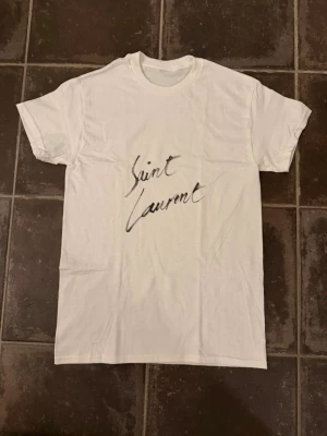 Saint Laurent t shirt  - Riktigt fet och eftertraktad t-shirt med Saint Laurent loggan i storlek S. Väldigt bra skick!  Obs: Jag är lite osäker på äktheten då lappen i nacken saknas, därför säljs den billigare (499 kr, nypris ca 3000 kr).  längd cirka 65cm, bredd cirka 44cm. Tveka inte att höra av dig vid frågor eller funderingar!