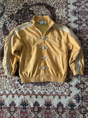 Gul Adidas jacka med vita detaljer - Unik gul Adidas jacka med vita ränder längs ärmarna och coola knytningar framtill. Jackan har ståkrage, två fickor och elastiska muddar. Ikoniska Adidas-loggan broderad på bröstet. Perfekt för dig som gillar retrostil och streetwear.