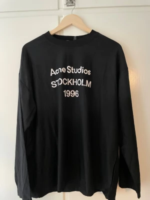 Svart långärmad topp Acne Studios - Svart långärmad med vit text 'Acne Studios Stockholm 1996' på bröstet. Klassisk rund halsringning och relaxed fit. Tillverkad i mjuk bomull, perfekt för en stilren och enkel look.
