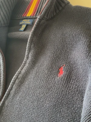 Grå stickad tröja från Polo Ralph Lauren - Mörkgrå stickad tröja från Polo Ralph Lauren med röd broderad logga på bröstet. Hög krage med ribbade detaljer och insida i rött och gult. Perfekt för dig som gillar klassisk och sportig stil. Priset går alltid att diskutera 