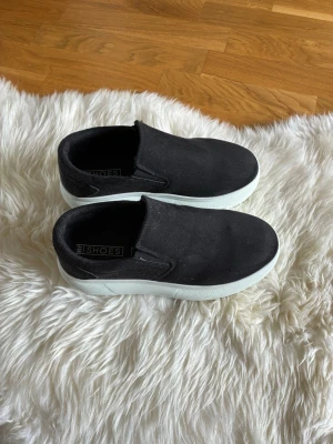 Svarta slip-on sneakers från My Shoes - Svarta slip-on sneakers från My Shoes med vit sula. Skorna har en enkel och stilren design utan snörning, vilket gör dem lätta att ta på. Ovandelen är i textilmaterial och sulan är i gummi. Perfekta för en avslappnad och trendig look.