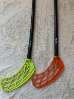 Innebandyklubbor och boll från Stiga - Två miniinnebandyklubbor med svarta skaft och blad i neonfärg, ett gult och ett orange. Till setet hör även en rosa innebandyboll med hål. Perfekt för innebandyspel hemma eller med kompisar.