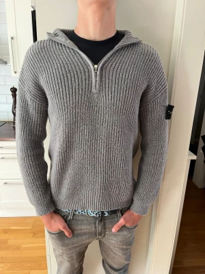 Grå stickad hoodie med dragkedja - Snygg grå stickad hoodie med halv dragkedja framtill och ribbstickad struktur. Tröjan har huva och en svart patch på ärmen som ger en cool detalj. Perfekt för lager-på-lager och chill vardagsstil.