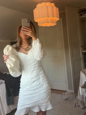 Vit kort klänning med volang - Supersöt vit kort klänning med v-ringning, långa puffärmar i transparent tyg och volangkant nertill. Klänningen har en figurnära passform och fina rynkade detaljer framtill. Perfekt för sommarens alla tillfällen.