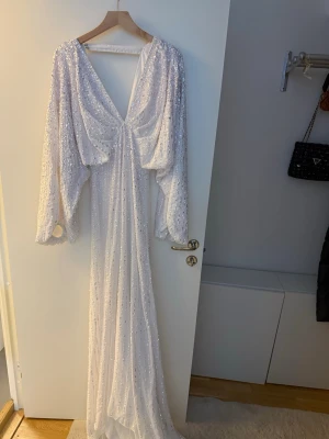 Asos bröllopsklänning  - Asos design slutsålda bröllopsklänning. Lennox vit långklänning med v-ringning och vida ärmar, täckt av glittriga paljetter. Klänningen har en elegant omlott-look framtill och markerad midja. Materialet känns lätt och skimrande. Endast testad