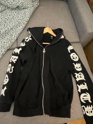 BlackSquad Hoodie - Svart hoodie med dragkedja och stora vita bokstäver i gotisk stil längs båda ärmarna och huvan. Hoodien har en klassisk passform, huva och ribbade muddar. Perfekt för dig som gillar streetwear och en edgy stil. Allt är perfekt, ej använt så många gånger. Är i mycket bra kondition. Passar för folk som gillar oversized clothes.