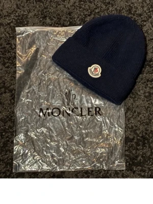 Mörkblå Moncler mössa i ull - Snygg mörkblå ribbstickad mössa från Moncler med klassisk logotyp framtill. Tillverkad i mjuk ull som håller dig varm och stilren under kalla dagar. Enkel, tidlös design som passar perfekt till streetwear och vinteroutfits.