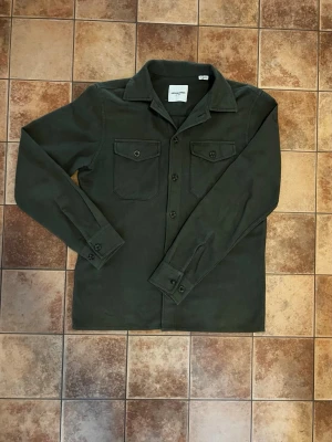 Olivgrön overshirt från Jack & Jones - Olivgrön overshirt från Jack & Jones i mjukt tyg. Klassisk krage, knäppning framtill och två bröstfickor med lock och knapp. Perfekt lager-på-lager-plagg med avslappnad passform och stilren look.