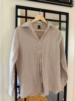 Randig skjorta från Only i linne - Snygg beige och vit randig skjorta från Only i luftigt linnematerial. Skjortan har klassisk krage, knäppning framtill och långa ärmar. Perfekt för en avslappnad och trendig look.