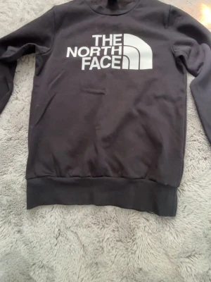 Svart sweatshirt från The North Face - Svart sweatshirt från The North Face med stor vit logga på bröstet. Tröjan har rund halsringning, långa ärmar och ribbade muddar vid ärmslut och nederkant. Perfekt för en avslappnad och stilren look.