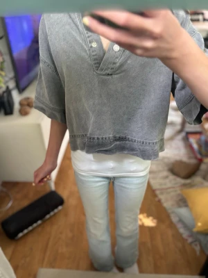 Grå croppad polotopp med knappar - Säljer en grå croppad polotopp med korta, vida ärmar och knappar vid halsen. Toppen har en avslappnad passform och är tillverkad i ett mjukt bomullsliknande material. Perfekt för en chill och trendig look.