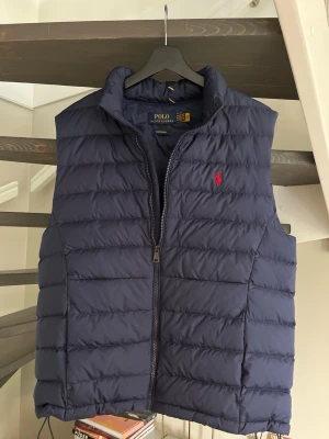 Mörkblå dunväst från Polo Ralph Lauren - Mörkblå Polo Ralph Lauren Performance väst med röd broderad logga och hög krage. Puffer-modell med dragkedja framtill. Bra skick.
