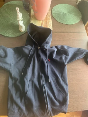 Mörkblå hoodie från Ralph Lauren - Snygg mörkblå hoodie med dragkedja från Ralph Lauren. Klassisk design med huva och dragsko, samt två fickor framtill. Liten röd logga broderad på bröstet. Perfekt för en chill och stilren look.