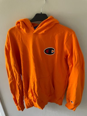 Orange hoodie från Champion - Snygg orange hoodie från Champion med stor logga på bröstet och liten logga på ärmen. Klassisk känguruficka framtill och skön huva. Perfekt för dig som vill sticka ut med färg och streetstyle.