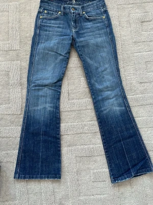 Blå bootcut jeans från 7 For All Mankind - Säljer ett par klassiska blå bootcut jeans från 7 For All Mankind. Jeansen har fem fickor, snygga slitningar och kontrastsömmar. Bakfickorna har det ikoniska A-broderiet. Tillverkade i mjuk denim med lätt stretch för skön passform.