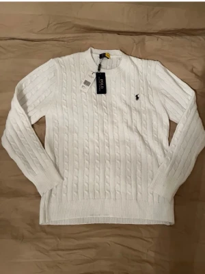 Vit stickad tröja Polo Ralph Lauren - Kabelstickad vit tröja från Polo Ralph Lauren med rund halsringning och svart broderad logga på bröstet. Tröjan har långa ärmar och ribbade muddar vid ärmslut och nederkant. Perfekt för dig som gillar klassisk och stilren design.